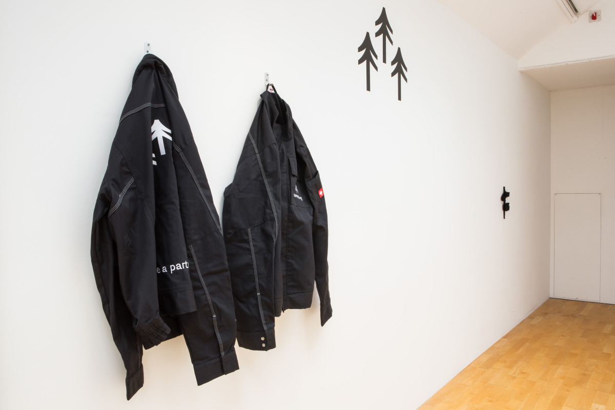 Two black work jackets on a wall with black trees vinyl on the wall, with small black woven sculpture on a white wall