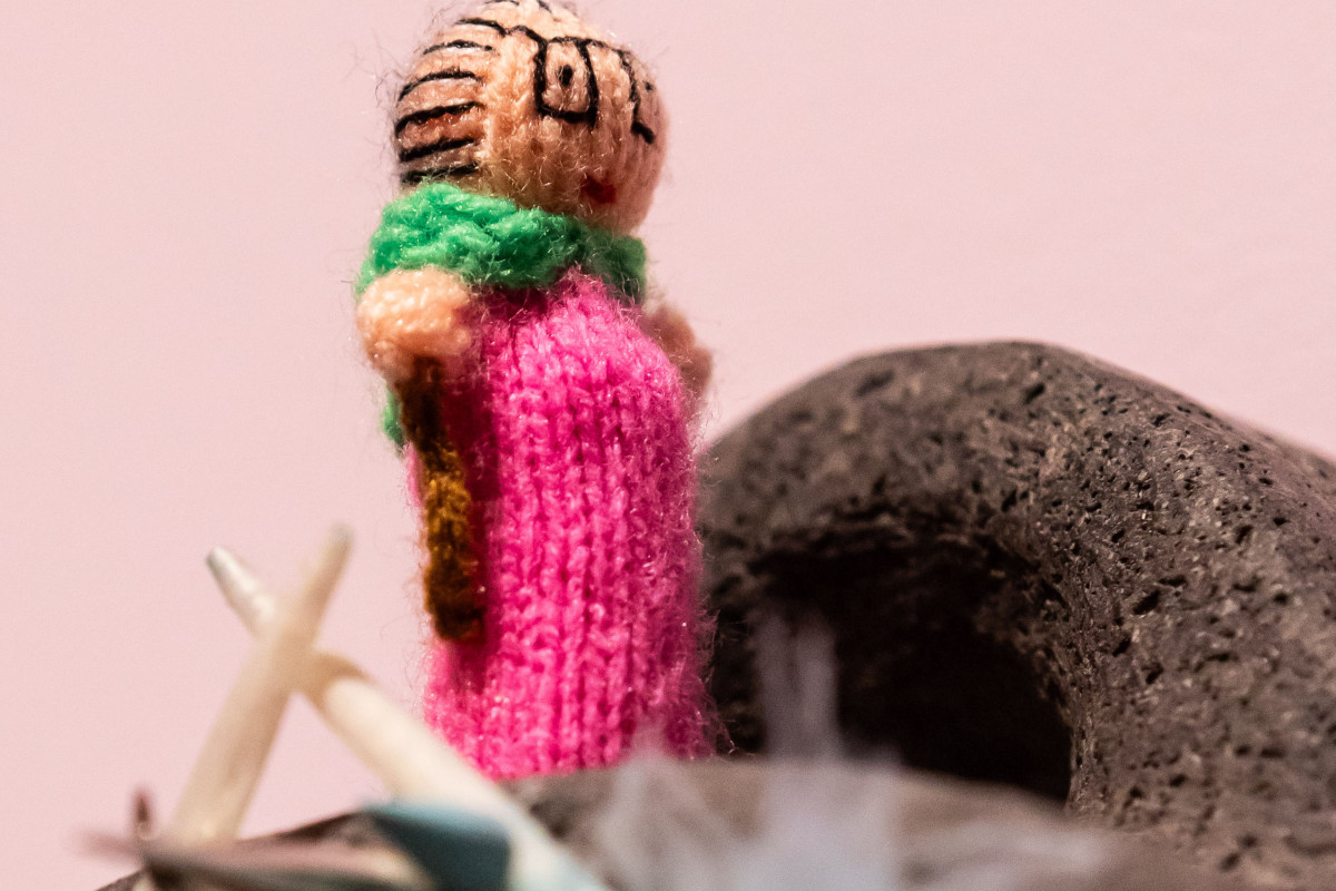 Fuzzy crochet figurine standing on volcanic rock and blue feathers against a pale pink background.